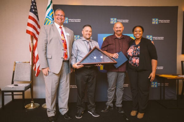 four individuals on stage with U.S. and NAfME flags and NAfME logo step and repeat backdrop. One individual holds framed folded U.S. flag that was flown over the U.S. Capitol building.