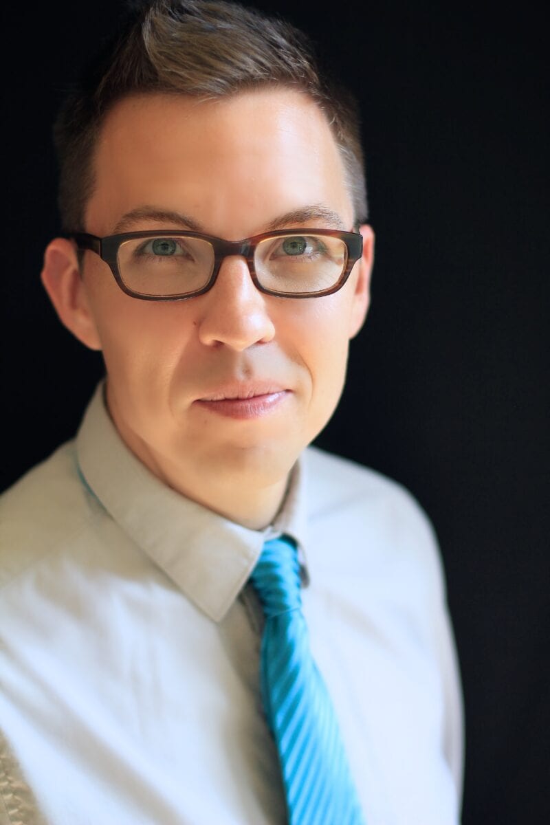 Joshua Palkki headshot wearing glasses, a white button down shirt, and blue diagonal striped tie