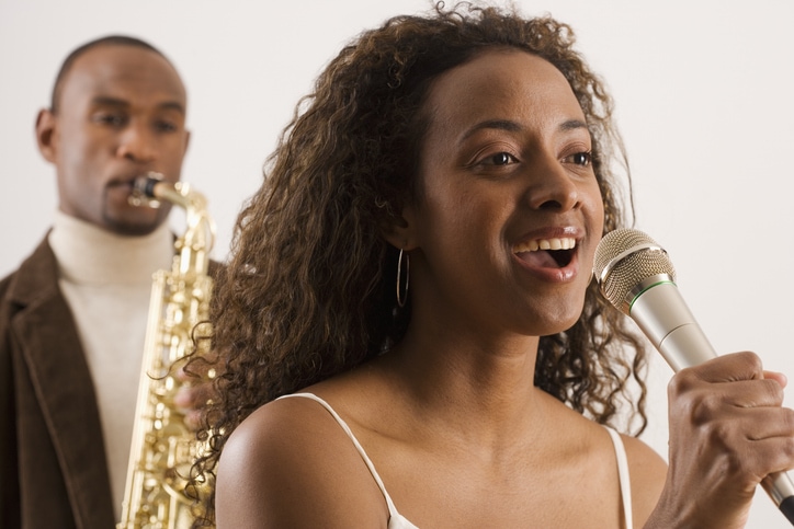 young woman singing into microphone with saxophonist playing behind her