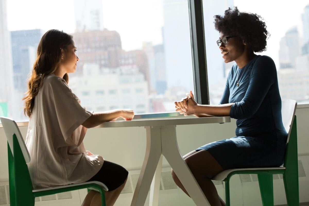 two women in conversation seated at a table by a window